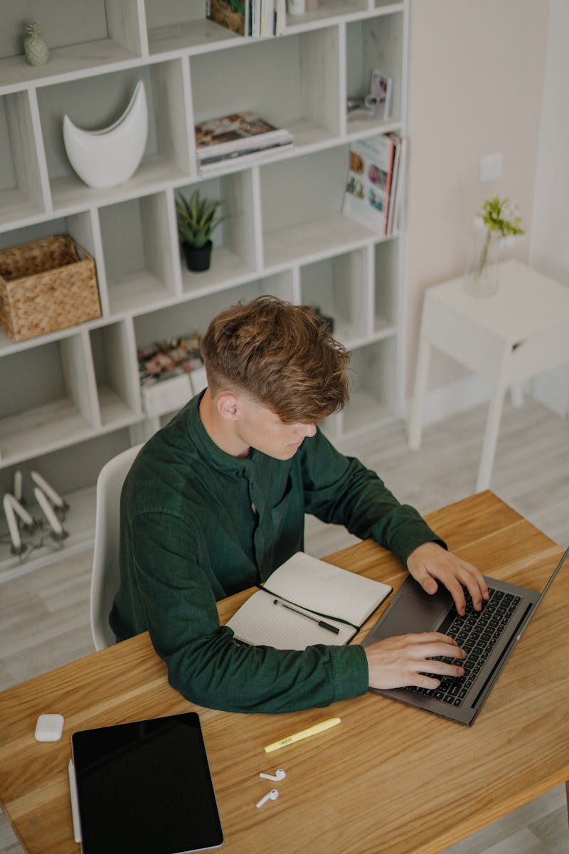 A young man using a laptop for distance learning at home, surrounded by modern gadgets on a wooden desk.